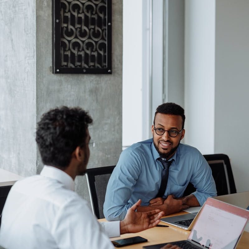 Two men in business attire discussing work at an office table with laptops.